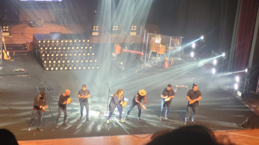 Foto do palco com Ana Carolina ao centro e três músicos de cada lado seu, com pandeiros nas mãos e fazendo uma performance musical.