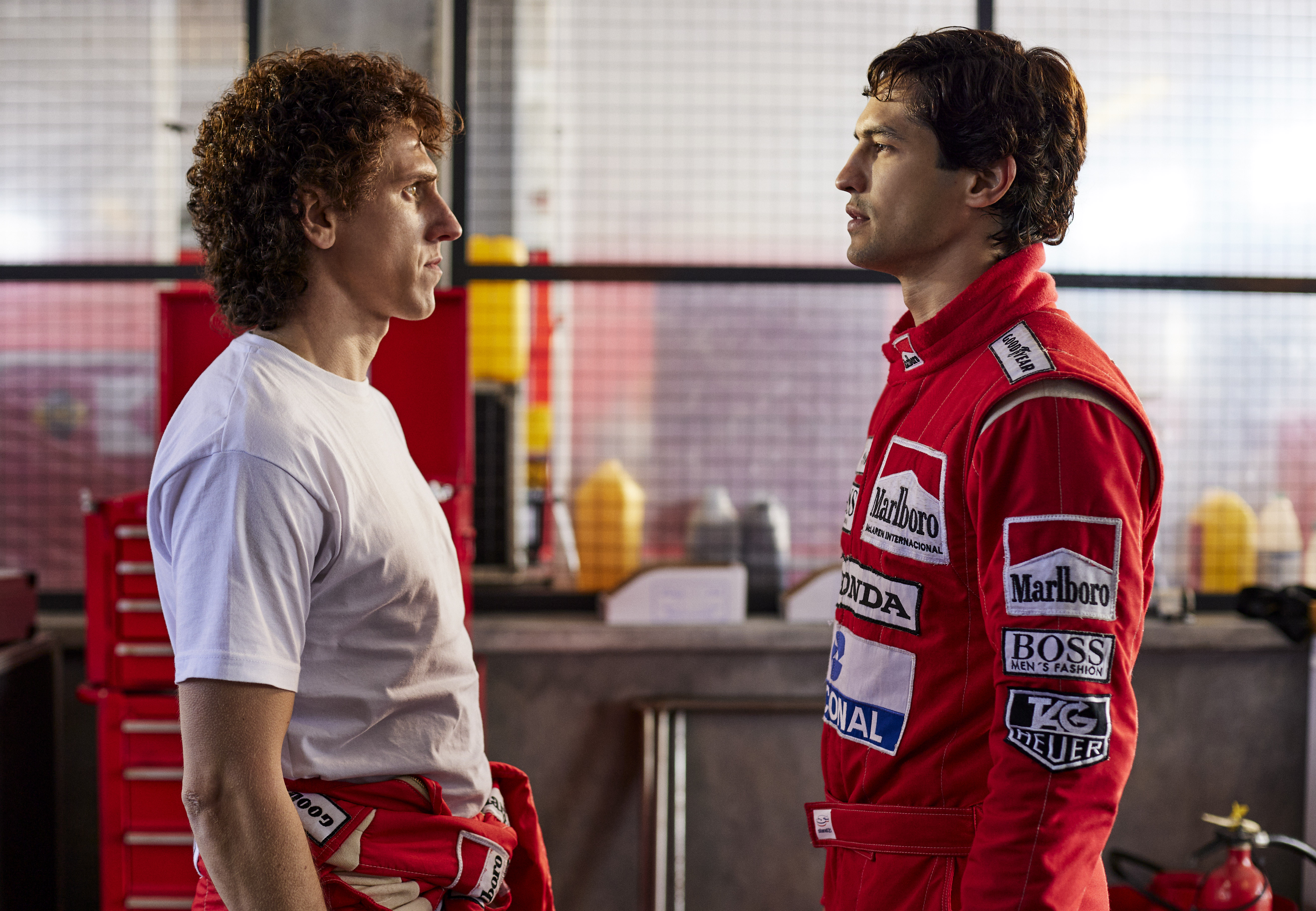Prost (Matt Mella), à esquerda, encarando Senna (Gabriel Leone), à direita. Prost está só de camiseta branca enquanto Senna veste o macacão vermelho da McLaren.