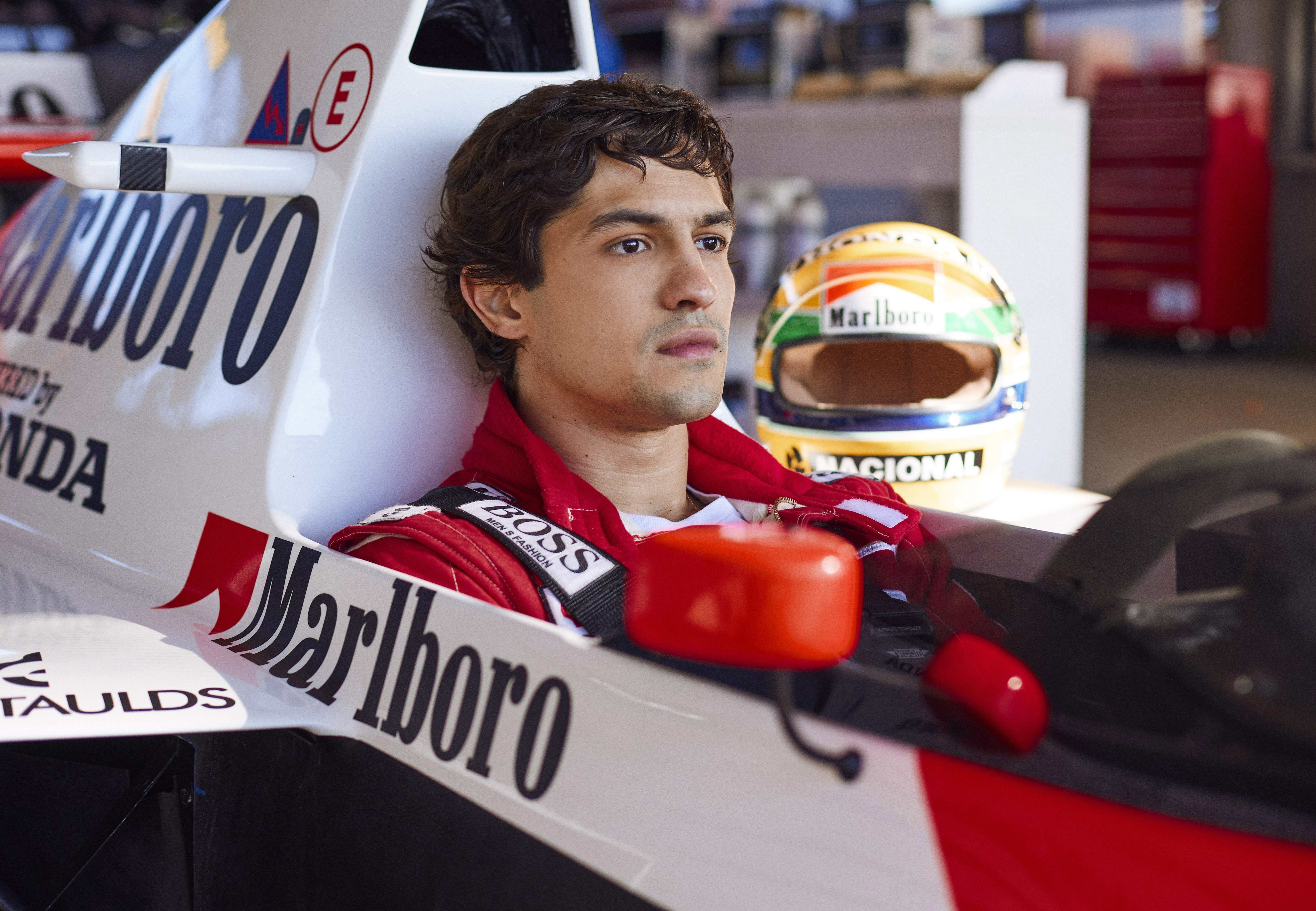 Senna (Gabriel Leone) sentado dentro de sua McLaren, com semblante pensativo e seu capacete repousando ao lado.
