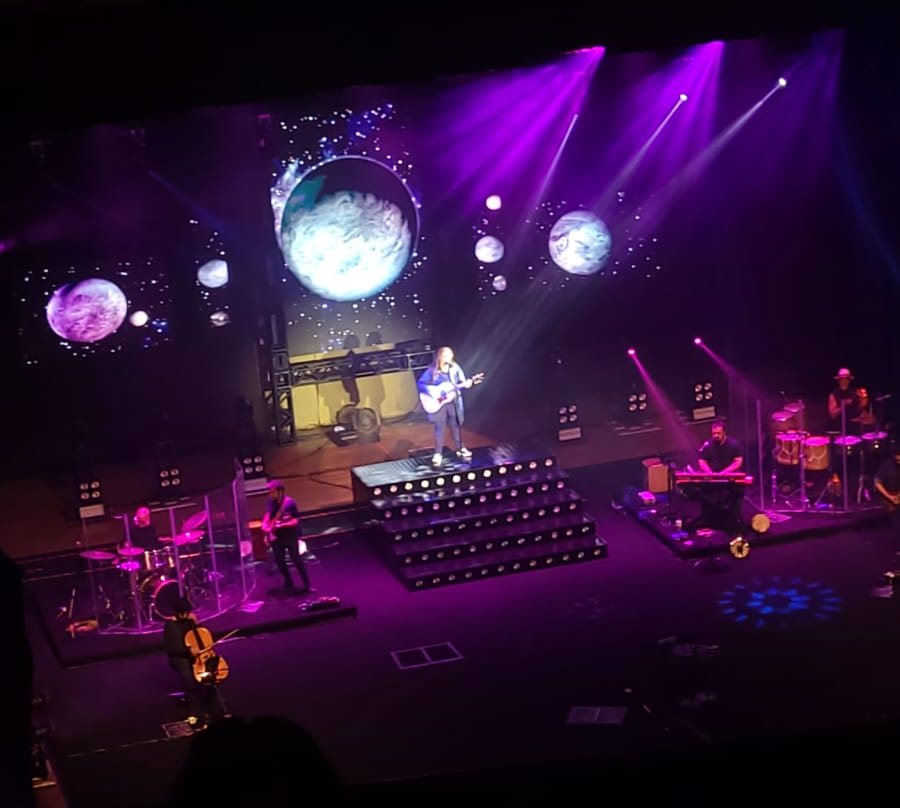 Ana ao centro, cantando e tocando violão, com toda a banda distribuída pelo palco, e no telão imagens de planetas e do espaço.