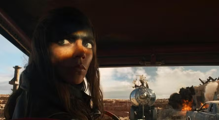 Cena do filme Furiosa com a protagonista ao volante, olhando para a câmera enquanto dá ré no carro, e uma explosão e brigas ocorrendo à frente, vistas pelo para brisas do veículo.