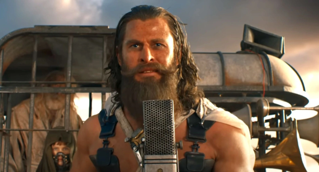 Dementus, personagem de Chris Hemsworth em Furiosa, encarando a cãmera com seus cabelos e barbas compridas, em frente a um microfone.