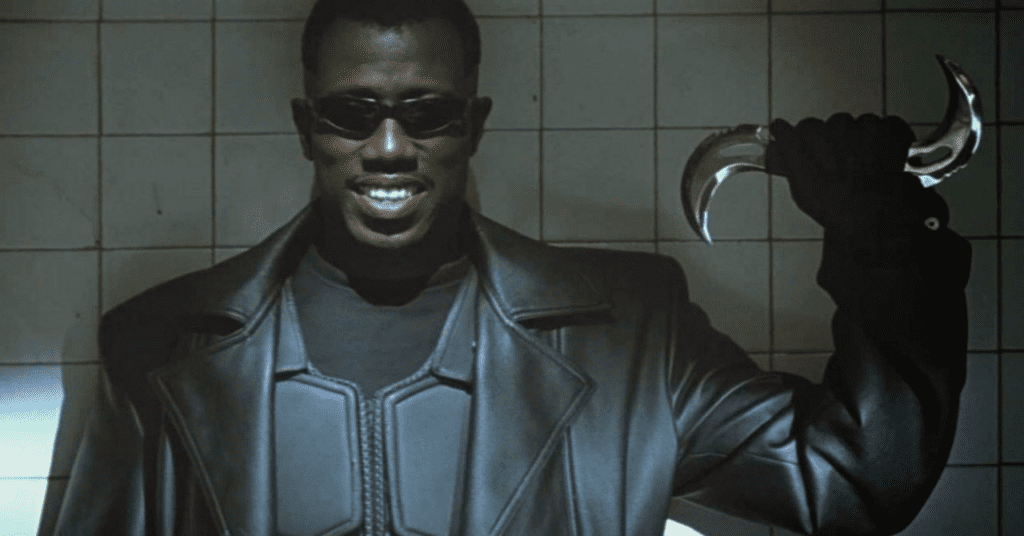 Wesley Snipes na pele de Blade, utilizando um sobretudo de couro e óculos escuros, segurando na mão esquerda uma espécie de bumerangue feito com duas lâminas curvadas, uma em cada ponta, em formato de S. Blade sorri para a câmera.