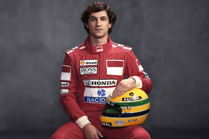 o ator Gabriel Leone, sentado em um banco, trajado com o macacão vermelho da McLaren e com o capacete amarelo com listras verde e azul em seu colo.