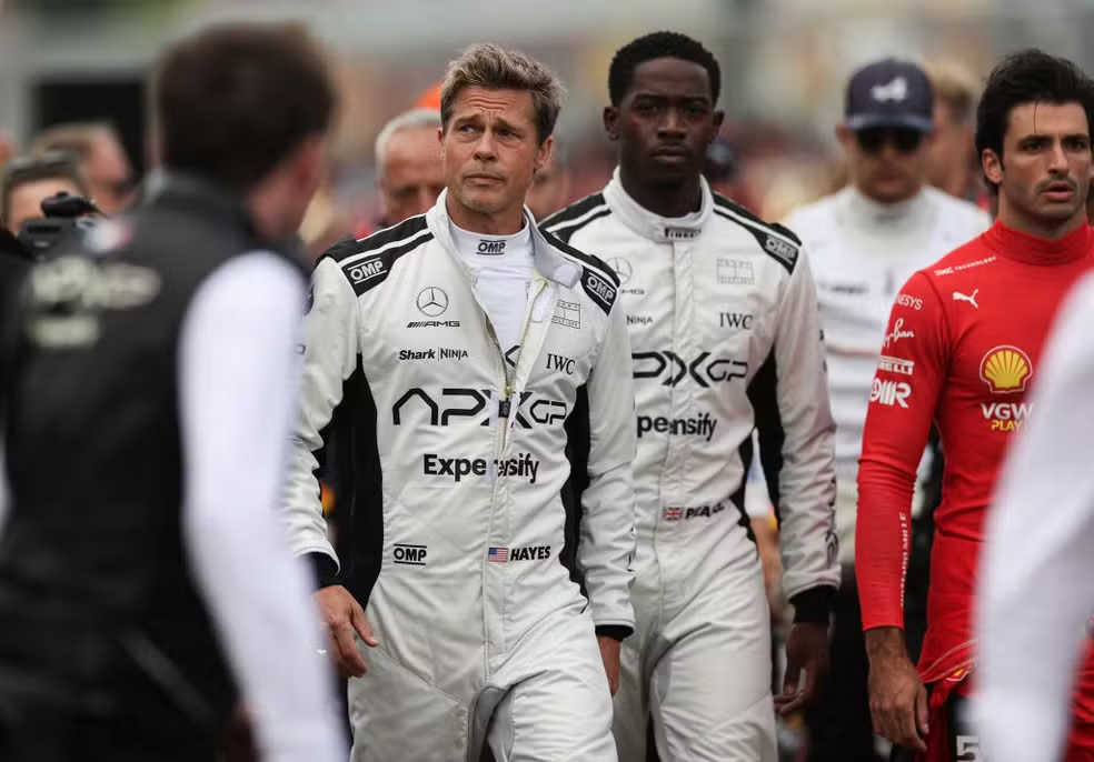 Brad Pitt e Damson Idris, trajados com o macacão da Apex GP (branco com patrocínios em preto) , e ao lado deles está Carlos Sainz, pilodo da Ferrari, também trajado com as vestimentas da escuderia.