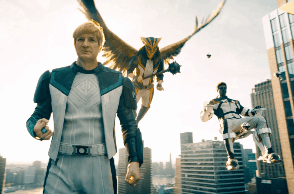 Nathan Fillion, de peruca loira, encarnando o herói Lanterna Verde, está em primeiro plano, com Mulher Gavião batendo asas e segurando uma maça logo atrás. Mais à direita, Sr Incrível está sentado em uma espécie de cadeira voadora. Todos estão devidamente trajados com uniformes de batalha branco e preto, com detalhes em verde e dourado.