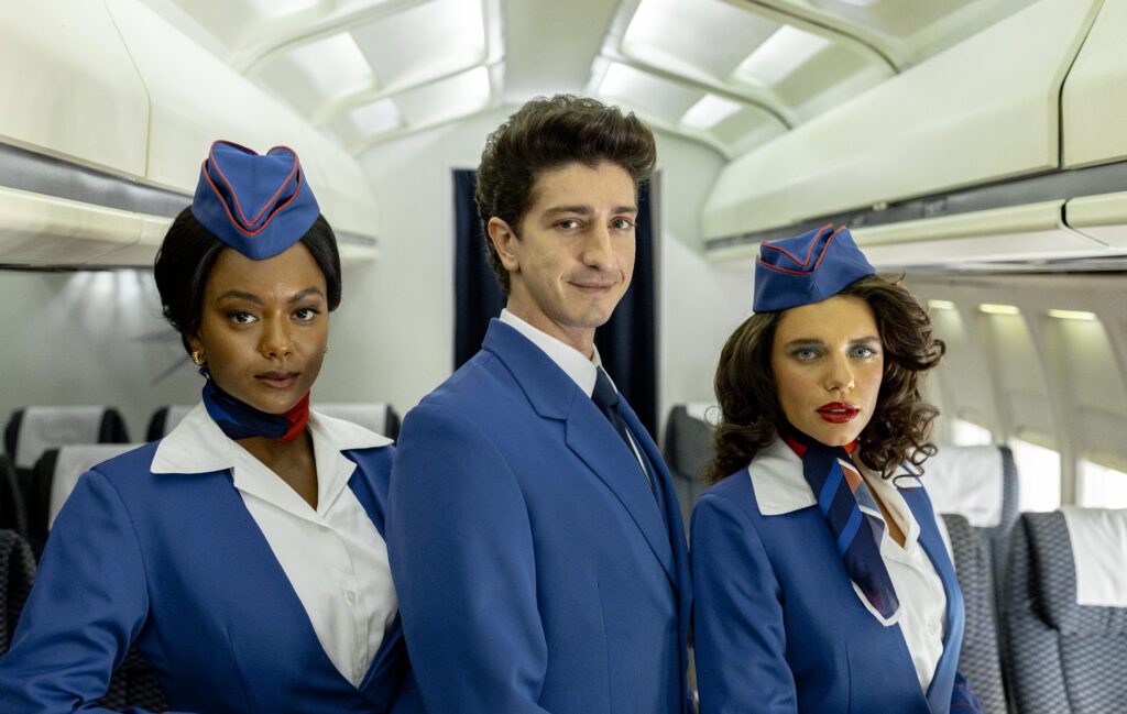 os atores Eli Ferreira, Johnny Massaro e Bruna Linzmeyer estão dentro de um avião, trajados como comissários de bordo em tons azuis, brancos e detalhes em vermelho.
