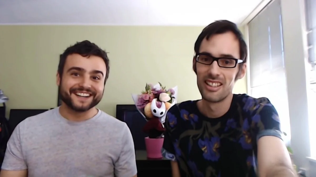 Os dois desenvolvedores olham para a tela durante uma chamada de vídeo. Estão vestidos informalmente, e ao fundo se vê um vaso de flores com uma pelúcia da personagem Hornet, protagonista do jogo Silksong.