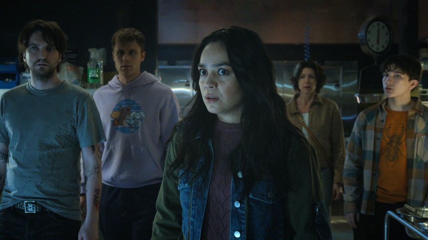 uma mulher de cabelos compridos e escuros encara com tensão no olhar algo que não é visto na imagem. Em segundo plano, mais quatro pessoas parecem olhar para a mesma coisa.