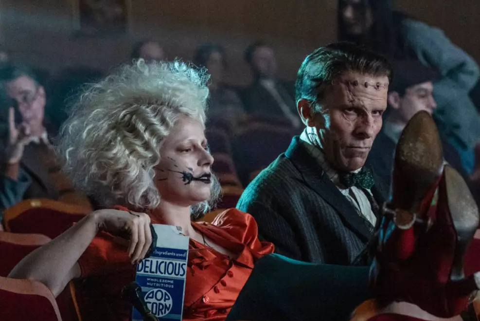 Penélope e Frank, sentados em poltronas de cinema. Penélope está com um pacote de pipocas na mão e as pernas cruzadas no banco da frente. Frank está de terno e gravata borboleta, encarando os pés de Penélope.