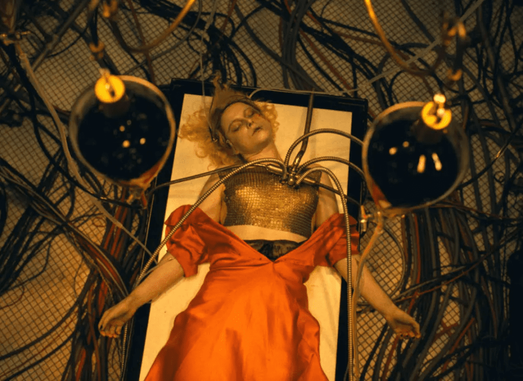 a protagonista está deitada em uma cama, cabeça tombada para a esquerda, com um vestido alaranjado semi aberto para comportar tubos que foram inseridos em seu tórax. Vários cabos de energia elétrica estão espalhados pelo chão.