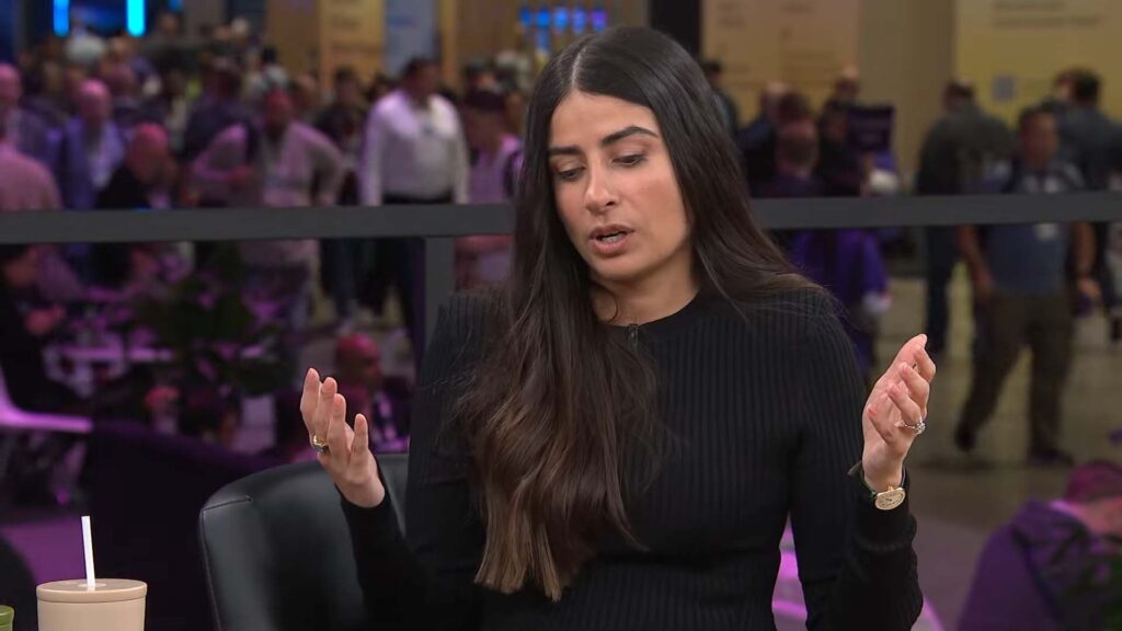 Ao centro, a CEO Asha Sharma. Ela é uma mulher branca de longos cabelos pretos e usa um suéter preto. Ela olha para baixo e suas mãos estão levantadas, a boca indica que fala algo no momento da foto.