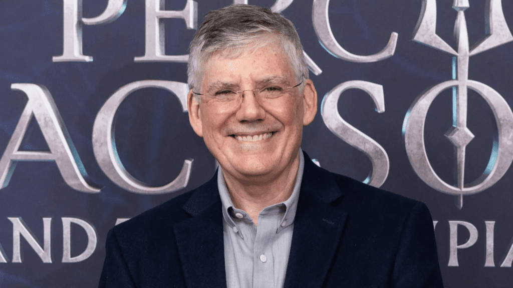 O autor Rick Riordan a frente e centralizado, ao fundo há a logo promocional de Percy Jackson. 