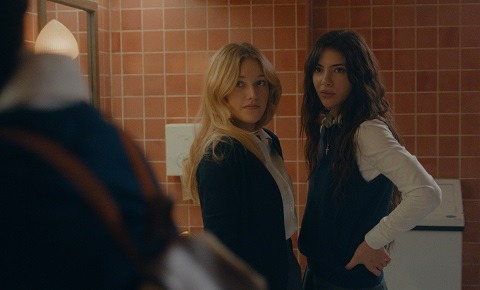 "Two young women in school uniforms stand in a tiled bathroom, looking towards someone entering. The background features a mirror, soft lighting, and a paper towel dispenser, creating a tense atmosphere."