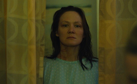 "A woman with wet hair and a hospital gown stands between yellow curtains, looking serious in a dimly lit room."