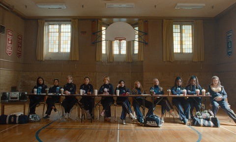 "A group of girls in athletic uniforms sit behind a long table in a gymnasium, with sports bags on the floor, under a basketball hoop and surrounded by tall windows and wall banners."