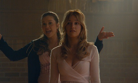 "Two young women stand in a warmly lit room with brick walls; one gestures and smiles energetically while the other looks ahead calmly, suggesting a lively conversation or instruction in a casual indoor setting."