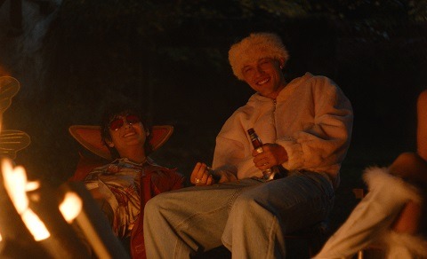 "Two people in costumes sit by a campfire at night, smiling and enjoying a relaxed outdoor setting; one holds a drink and both wear warm, playful outfits."