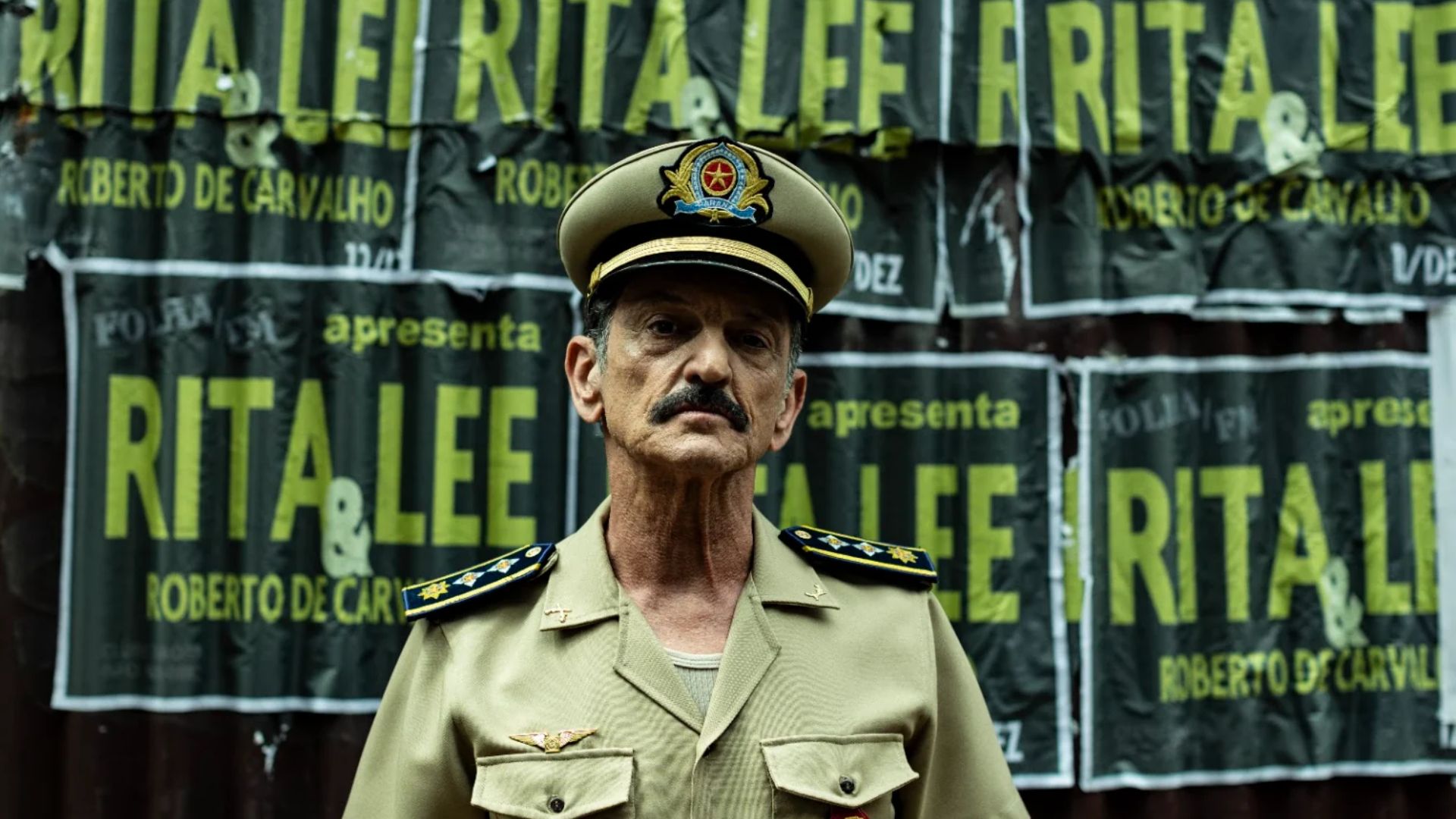 Cena de Assalto à Brasileira com um oficial da polícia militar diante de cartazes de show da Rita Lee